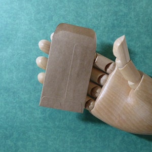 May include: A brown paper envelope with a flap on the top. The envelope is held in a wooden hand.
