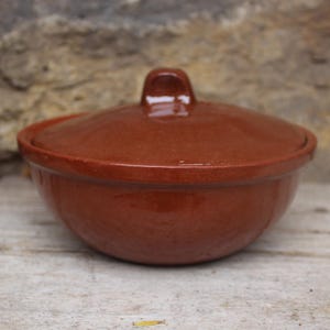 Op de afbeelding: Een kleine, ronde keramische kom met deksel in een warme, roodbruine tint. Het deksel heeft een kleine, rechthoekige handgreep. De kom staat op een verweerd houten oppervlak, wat wijst op een rustieke of handgemaakte uitstraling. De kom wordt waarschijnlijk gebruikt voor het bewaren of serveren van voedsel.