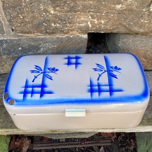 May include: A vintage, rectangular, beige and blue container. The lid is light beige with a blue border and floral and grid designs. The container has a white clasp and is sitting on a wooden surface.