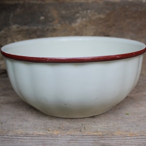 May include: White enamel bowl with a red rim. The bowl has a fluted design and is sitting on a wooden surface.