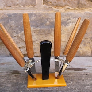 Könnte beinhalten: Ein Set aus sechs Vintage-Buttermessern mit Holzgriffen und verchromten Klingen. Die Messer sind fächerförmig in einem schwarzen und gelben Kunststoffständer angeordnet.
