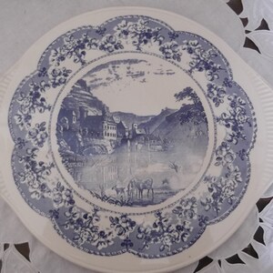 Könnte beinhalten: Ein weißer Keramikteller mit einem blauen und weißen Blumendesign. Die Mitte des Tellers zeigt eine blaue und weiße Landschaftsszene mit einem Dorf und einem Fluss.
