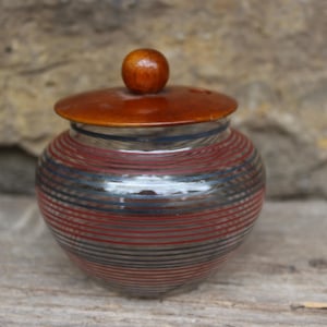 Peut inclure: Petit pot rond avec couvercle et bouton en bois. Le pot est orné de rayures horizontales rouges, bleues et noires. Le couvercle est de couleur marron chaud et le bouton est marron foncé. Le pot est posé sur une surface en bois.