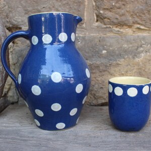 Könnte beinhalten: Eine blaue Keramikkanne und eine passende blaue Keramiktasse mit weißen Punkten. Die Kanne hat einen Henkel und einen Ausguss. Die Tasse hat eine Standardgröße.