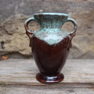 Könnte beinhalten: Eine braune Keramikvase mit zwei Henkeln und einem ausgestellten Rand. Die Oberseite der Vase ist eine gesprenkelte türkisfarbene Farbe, die in einen glänzenden braunen Körper übergeht. Die Vase hat einen abgerundeten Boden und dient wahrscheinlich dekorativen Zwecken.