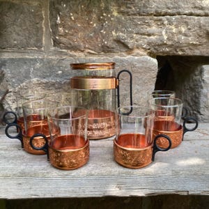 May include: A glass pitcher and four matching glass cups, each featuring a textured copper-colored metal base and a black handle. The pitcher has a copper-colored metal band around the top and a black handle, completing this drinkware set.