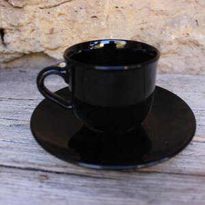 Op de afbeelding: Een glanzende zwarte espressokop en schotel set. De kop heeft een gebogen oor en staat op een bijpassende ronde schotel. De set is gemaakt van een reflecterend materiaal en de donkere kleur geeft het een strakke, moderne uitstraling. De set staat op een houten oppervlak.