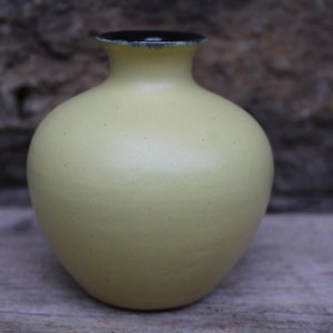 May include: A small, round, yellow ceramic vase with a black rim. The vase is sitting on a wooden surface.