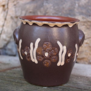 May include: A brown ceramic pot with white and brown floral designs. The pot has two small handles on the sides.