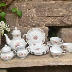 May include: A white porcelain tea set with a floral design. The set includes a teapot, sugar bowl, creamer, plates, and teacups. The floral pattern features purple and yellow flowers with green leaves. The set is arranged on a wooden surface.