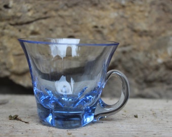 May include: A small, clear glass cup with a blue rim and base. The cup has a handle and a flared lip.