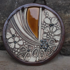 Könnte beinhalten: Ein runder Wandbehang aus Keramik mit braunem Rand und weißen Blumenmustern. Die Mitte des Wandbehangs hat einen braunen und orangefarbenen glasierten Bereich.