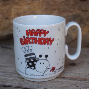 May include: White ceramic mug with a handle, featuring the words "HAPPY BIRTHDAY" in red. The mug has a cartoon illustration of a smiling bug with a birthday cake on its back. The mug is on a wooden surface.