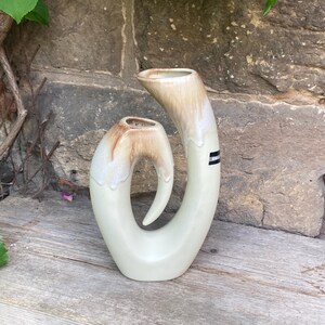 Könnte beinhalten: Eine Keramikvase mit zwei Öffnungen, einer glatten, cremeweißen Basis und einer glänzenden, braunen Tropfglasur an den oberen Rändern. Die Struktur besteht aus zwei miteinander verbundenen, geschwungenen, hornartigen Ausläufen mit einem kleinen schwarz-weißen Etikett.