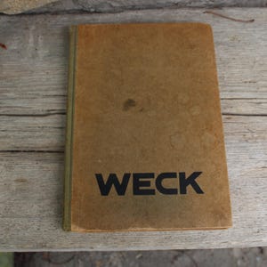May include: A vintage, tan-coloured book with the word "WECK" printed in bold black letters. The book has a slightly worn appearance, with a few dark spots on the cover. The book is resting on a weathered wooden surface.