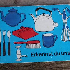 Può includere: Una carta illustrata con una varietà di oggetti per la casa, tra cui una teiera, un bollitore, una paletta per la polvere, una spazzola, una tazza, un rubinetto e una frusta. Il testo sulla carta dice "Erkennst du uns".