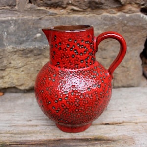Puede incluir: Una jarra de cerámica roja con cuerpo bulboso, pico vertedor y asa curva. La jarra tiene una superficie texturizada con pequeños puntos oscuros. El interior de la jarra es marrón. La jarra está sobre una superficie de madera.