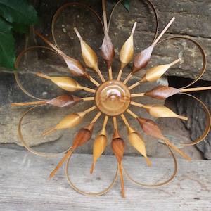 Op de afbeelding: Houten wandkunst in een bloemmotief. Het midden is een lichtbruine cirkel met stralende houten elementen in bruin- en beigetinten. De buitenste bloembladen zijn gebogen, dun en gemaakt van lichtbruin materiaal.