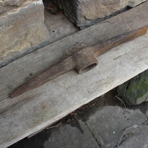 Op de afbeelding: Een roestige metalen pickaxe met een houten steel. De pickaxe ligt op een verweerd houten plank.