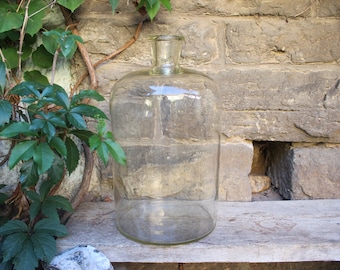 May include: A large, clear glass carboy with a wide, cylindrical body and a narrow, short neck. The vessel features a smooth, transparent surface and a thick, rounded rim at the top opening, designed for storage or fermentation purposes.