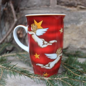 Puede incluir: Taza de cerámica roja con asa blanca, con ilustraciones de ángeles y estrellas. La taza tiene un acabado brillante y está decorada con estrellas doradas y figuras angelicales con alas blancas. Un artículo decorativo festivo.