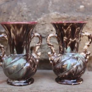 Puede incluir: Dos jarrones de cerámica marrón y verde con asas ornamentadas. Los jarrones tienen un esmalte marmoleado y un borde rojo.