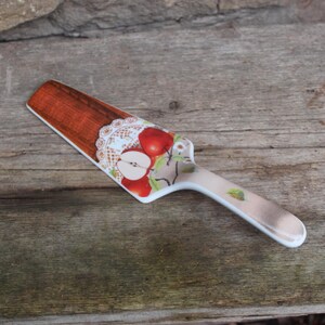 Może przedstawiać: Biały ceramiczny nóż do ciasta z brązowym i białym wzorem przedstawiającym jabłka i koronkę. Nóż ma drewniany uchwyt.