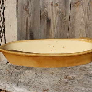 Pode incluir: Uma assadeira de cerâmica ovalada, com interior amarelo claro e exterior castanho quente. A assadeira está sobre uma superfície de madeira desgastada, em frente a um fundo de madeira rústica. A assadeira tem cerca de 36 cm de comprimento.