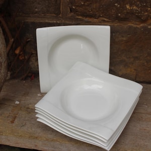 Puede incluir: Un conjunto de cuencos blancos cuadrados para sopa con un centro redondeado. Los cuencos de porcelana tienen un diseño sencillo y elegante. Los cuencos están apilados sobre una superficie de madera.