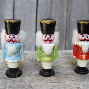 May include: Three wooden nutcracker figurines with painted faces and white fur beards. The nutcrackers are wearing blue, green, and red uniforms with gold accents.