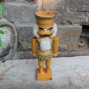 Könnte beinhalten: Ein Nussknacker aus Holz mit weißem Bart und goldener Verzierung um den Hut. Der Nussknacker hat ein bemaltes Gesicht mit einem Schnurrbart und Augenbrauen.