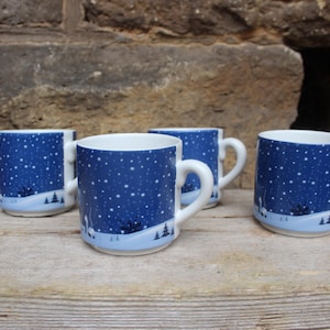 Puede incluir: Cuatro tazas de cerámica con un diseño de escena invernal. Las tazas tienen un interior blanco y un exterior azul oscuro con estrellas blancas y un paisaje invernal. Cada taza tiene un asa blanca y una base azul claro.