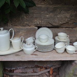 May include: A complete cream-colored tea set with silver trim, including a teapot, creamer, sugar bowl, serving tray, plates, and teacups. The set is arranged on a wooden surface against a stone wall, ideal for serving tea.
