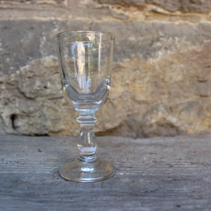 May include: A clear glass shot glass with a stemmed base. The glass is empty and sits on a wooden surface.