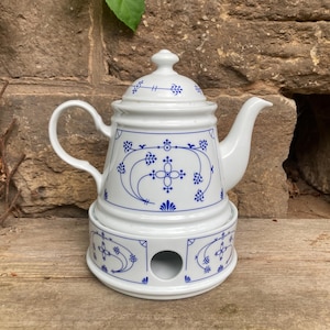 Op de afbeelding: Witte keramische theepot met een bijpassende warmhoudbasis, beide versierd met blauwe bloemen en geometrische patronen. De theepot heeft een gebogen handvat en een tuit. De warmhoudbasis heeft een cirkelvormige opening. De theepot staat op een houten oppervlak.