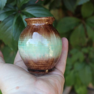 May include: A small, round ceramic vase with a brown and green glaze. The vase is approximately 7.5 cm tall and has a wide mouth.