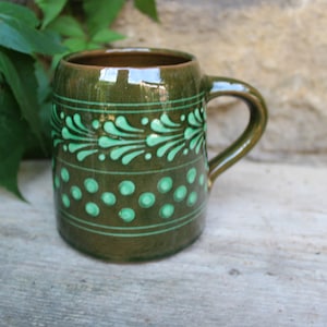 Könnte beinhalten: Eine grüne Keramiktasse mit einem grün-weißen Blumendesign. Die Tasse hat einen Henkel und steht auf einer Holzoberfläche.