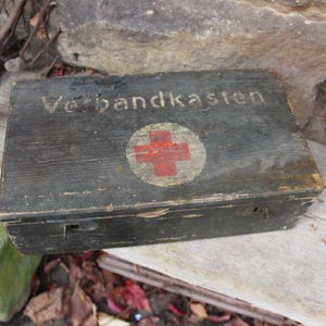 May include: A dark, weathered wooden box, likely a vintage first aid kit, with the German text "Verbandkasten" and a red cross emblem. The rectangular box has a hinged lid and a latch. The box is made of dark wood.