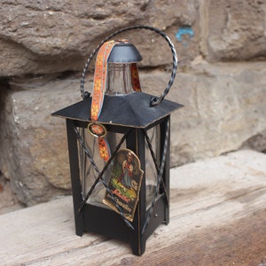 May include: Black metal lantern with a glass bottle inside. The bottle has a label that says "Lebertran" and a picture of a woman in a traditional dress. The lantern has a handle and a decorative ribbon.