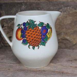 May include: White ceramic jug with a fruit design. The design features a pineapple, banana, plums, and berries in shades of orange, yellow, blue, and green.