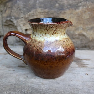 May include: A small brown and cream ceramic jug with a speckled glaze. The jug has a rounded body and a handle.