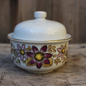 Könnte beinhalten: Weiße Zuckerdose aus Keramik mit Deckel, verziert mit einem Blumendesign mit rosa, lila und gelben Blumen auf weißem Hintergrund mit goldenen Akzenten.