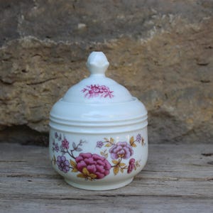 Puede incluir: Un azucarero de cerámica blanca con tapa, adornado con un estampado floral en tonos rosa, morado y amarillo. El azucarero tiene forma redondeada y está sobre una superficie de madera, con un fondo de piedra.