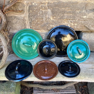 Può includere: Una collezione di coperchi per pentole in smalto vintage di varie dimensioni e colori, tra cui verde, nero e marrone. I coperchi sono disposti su una superficie di legno, mostrando le loro diverse forme e design delle maniglie. I coperchi provengono probabilmente da un set di pentole.