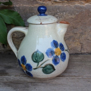 Può includere: Un piccolo brocca in ceramica bianca con un motivo floreale blu e giallo. La brocca ha un coperchio blu con un pomo in cima.