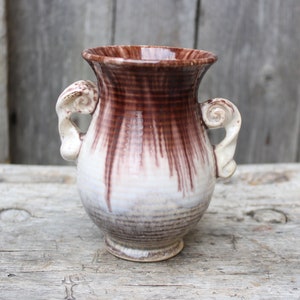 Puede incluir: Un pequeño jarrón de cerámica blanca con esmalte marrón y dos asas decorativas. El jarrón tiene un cuerpo redondeado y un cuello estrecho.