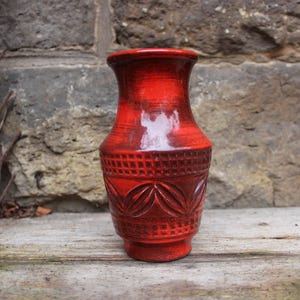 May include: A red ceramic vase with a textured surface and a flared rim. The vase features a patterned design around the middle and is approximately 20 cm tall. The vase is a decorative item.