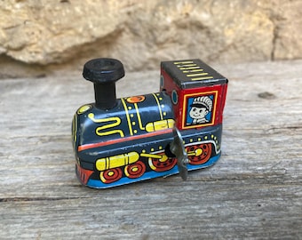 Könnte beinhalten: Eine Spielzeug-Dampflokomotive aus Blech im Vintage-Stil mit dunkelblauem Gehäuse, gelben und roten Farbakzenten, einem schwarzen Schornstein sowie einem roten Führerhaus, das ein illustriertes Gesicht mit gestreifter Mütze im Fenster zeigt.