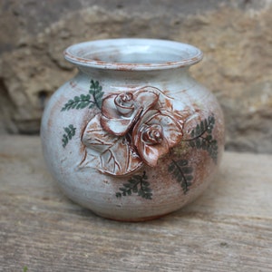 Könnte beinhalten: Eine weiße Keramikvase mit brauner Glasur und einem erhabenen Blumendesign. Die Vase ist mit zwei Rosen und Farnblättern verziert.