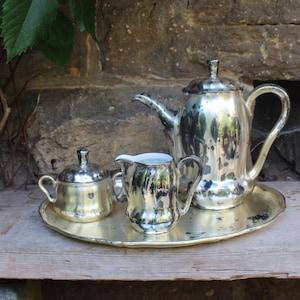 Puede incluir: Un juego de té plateado vintage, que incluye una tetera, una jarra de crema, un azucarero y una bandeja redonda. El juego se exhibe sobre una superficie de madera contra una pared de piedra. El juego de té es de diseño clásico.